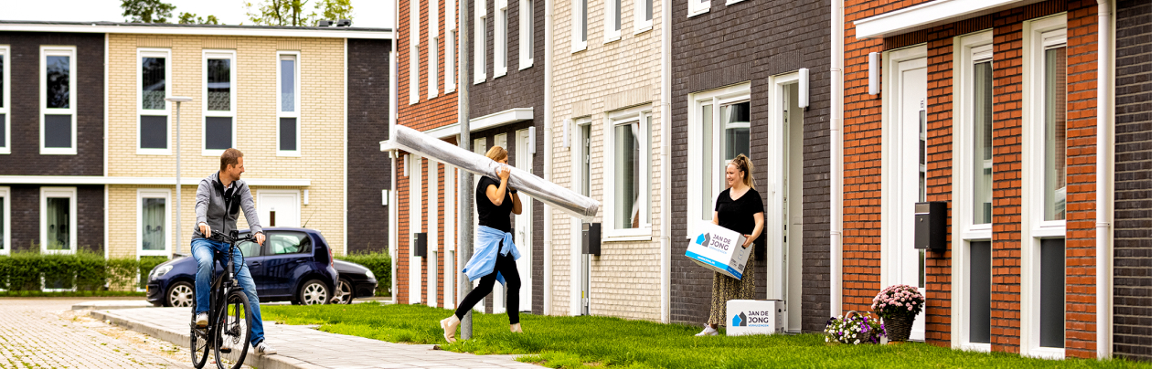 Zwei Frauen tragen Kartons und einen eingerollten Teppich in ein Reihenhaus. Ein Mann auf einem Fahrrad fährt daran vorbei.