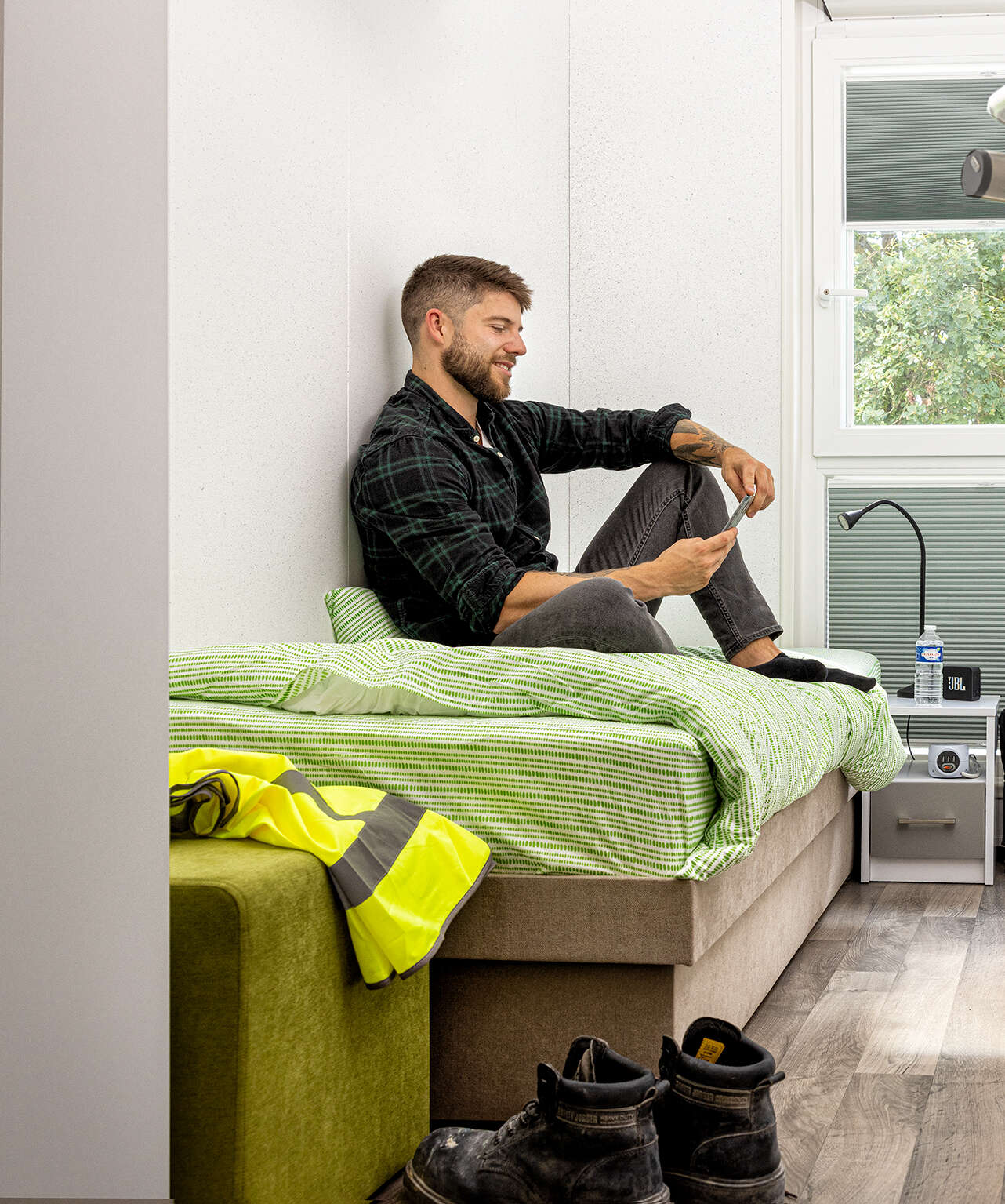 Einblick Schlafzimmer Wohngemeinschaft. Auf dem Bett sieht man einen Arbeiter sitzen, der auf sein Handy schaut.