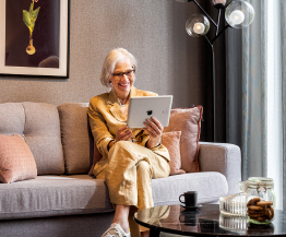 Dame sitzt mit Tablet auf Sofa einer Seniorenwohnung